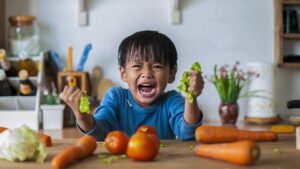 A child sits at a table of vegetables and screams, displaying aggressive behavior.