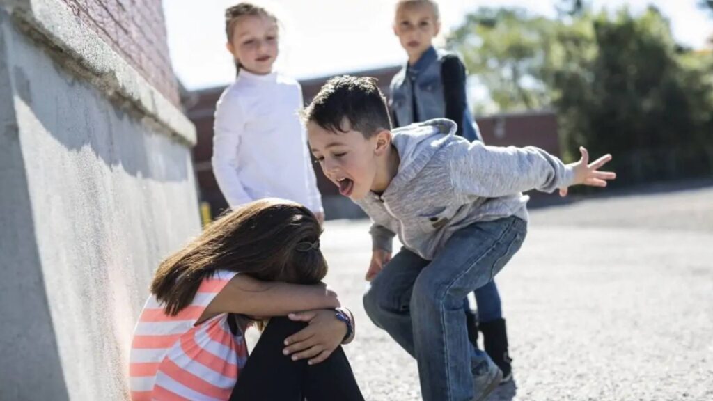 A child stands in front of another child and bullies her. Two bystanders stand in the background, watching.