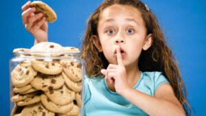 A kid sneaks a cookie from the cookie jar and is holding her finger to her lip to say "shh." She is demonstrating the need for kids to eat healthy.