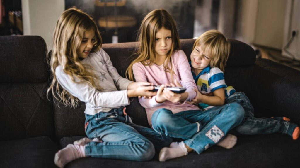 Three kids sit on a black couch in front of a television, fighting over the remote control, demonstrating screen addiction.